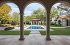 View from back patio with archway, columns, and tiled floor, overlooking square pool with custom fountain, pool-house and gorgeous landscaping